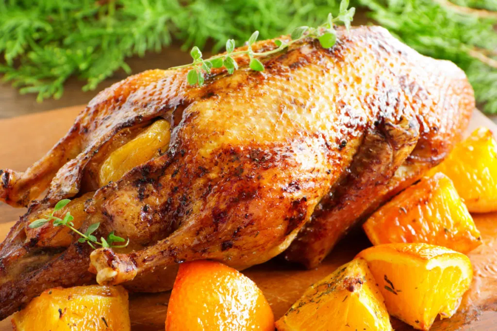 A roast duck with oranges arranged on a cutting board, there are orange slices next to the crispy bird. There are nondescript herbs in the background.
