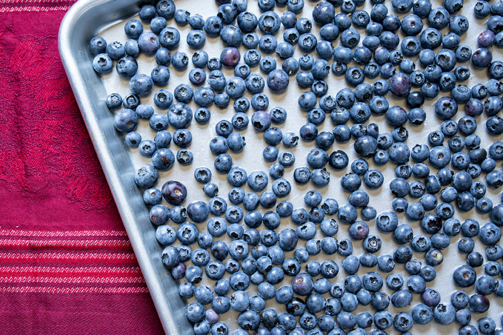 Overhead view of a half-sheet baking pan. There are fresh blueberries lining the pan in a single layer. The pan is sitting on a maroon kitchen towel at an angle.
