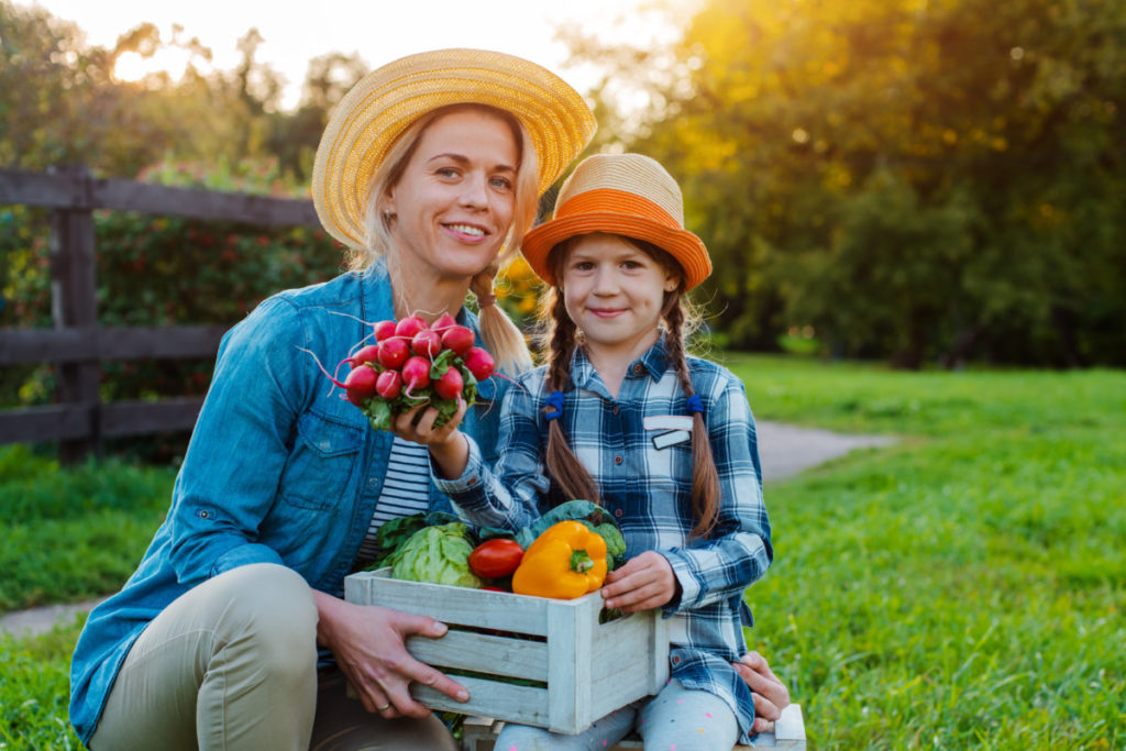 A smiling mother and daughter in tidy, clean outfits smile while holding a box of freshly picked, shiny produce. The daughter is holding up a bunch of radishes. The sun is setting behind the trees behind them both.