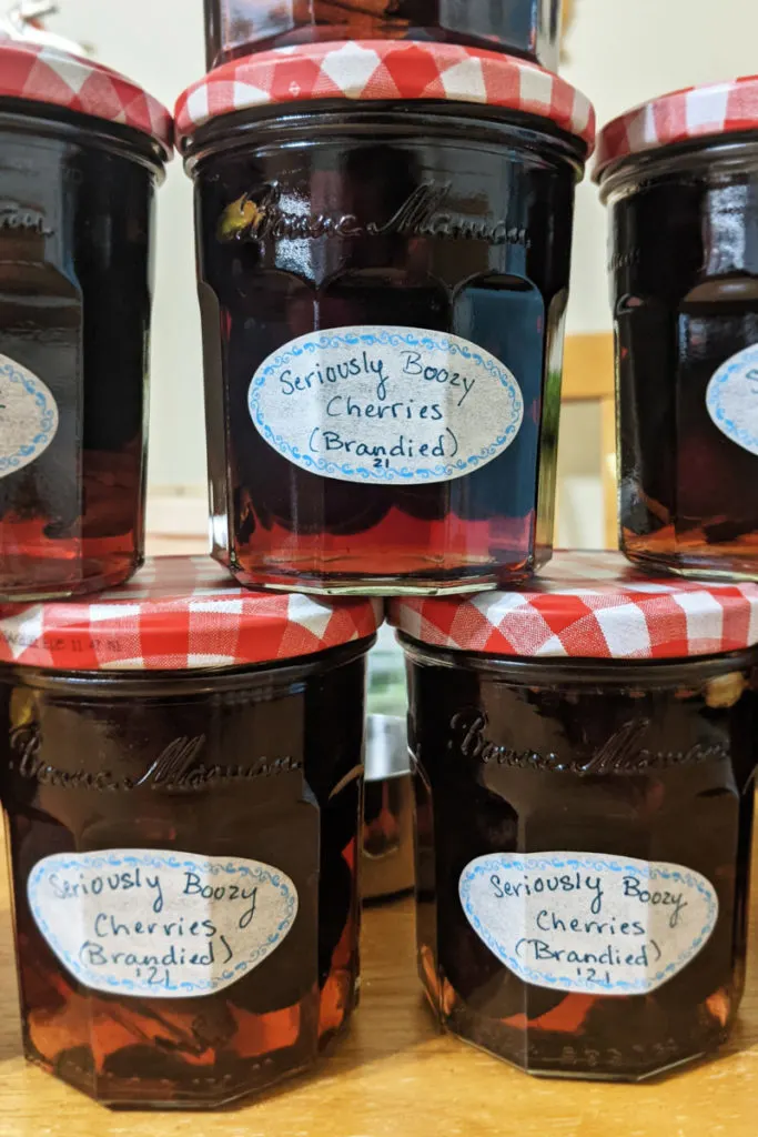The same jars now filled with the brandy and honey syrup, with their lids on them, and stacked one on top of the other. The lids are a red and white checked gingham and the jars have been labeled "Seriously Boozy Cherries (Brandied) '21"