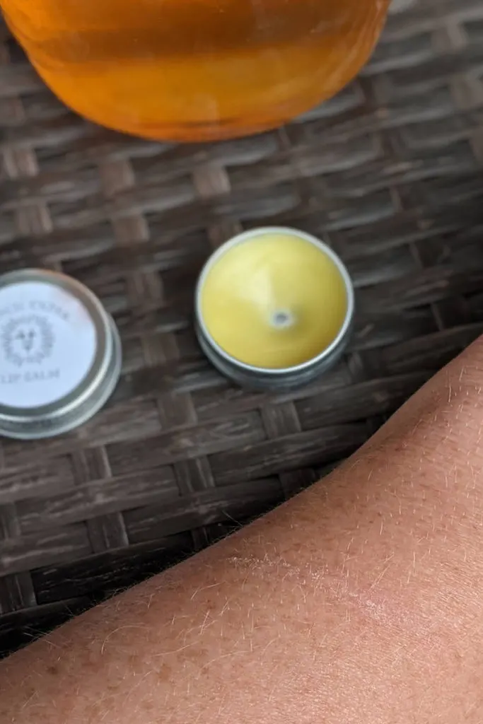 The same tin of lip balm on a rattan table. Someone's arm is next to it with a small scratch. In the background you can see the bottom of a jar of honey.