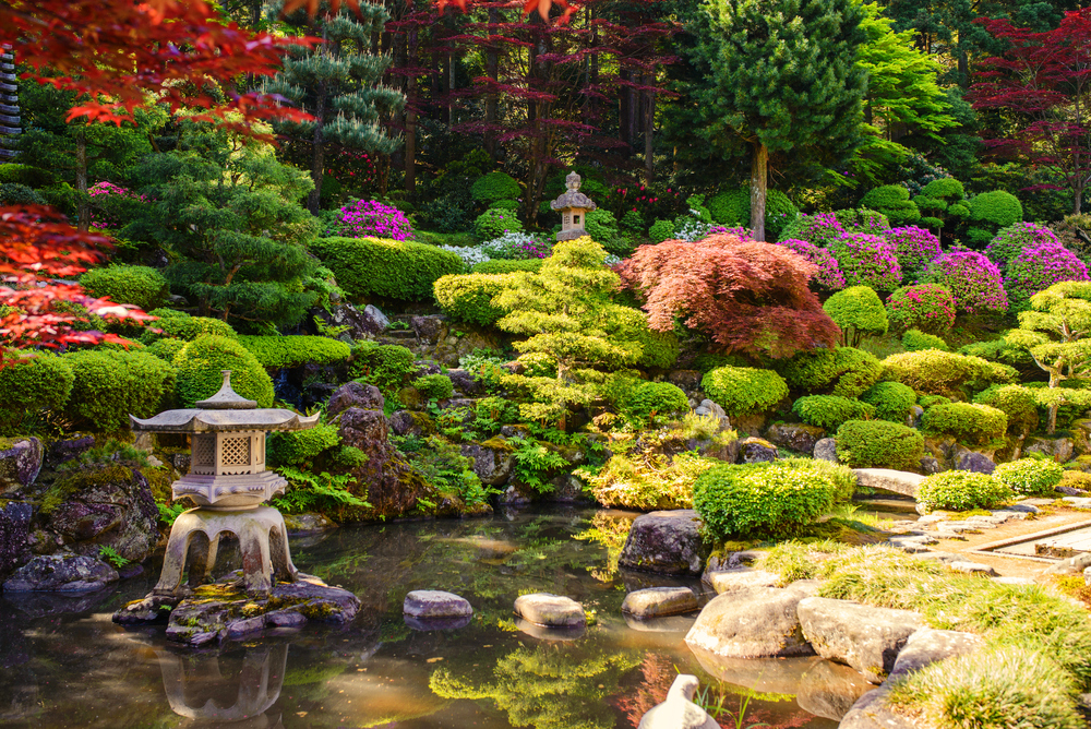 A lush and colorful Japanese pond with sculpted shrubs and flowers growing. There is a small cement pagoda statue standing in the pond.