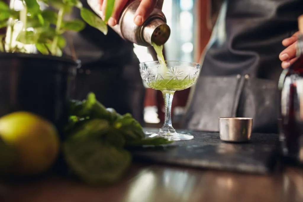 A bartender pours a green cocktail into a chilled coupe glass.
