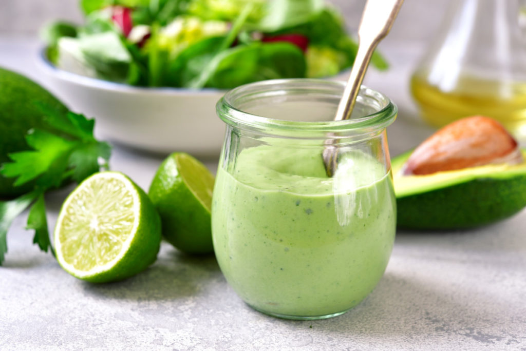 A brightly lit scene. A small clear round jar filled with bright green dressing with a spoon in it. Behind is a sliced avocado, a sliced lime and a salad.