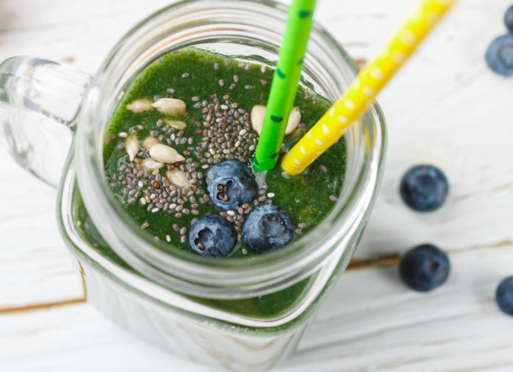 Close up overhead view of kale smoothie in a mason jar mug. The smoothie is topped with sunflower seeds, chia seeds, and blueberries. There are two paper straws in the smoothie.