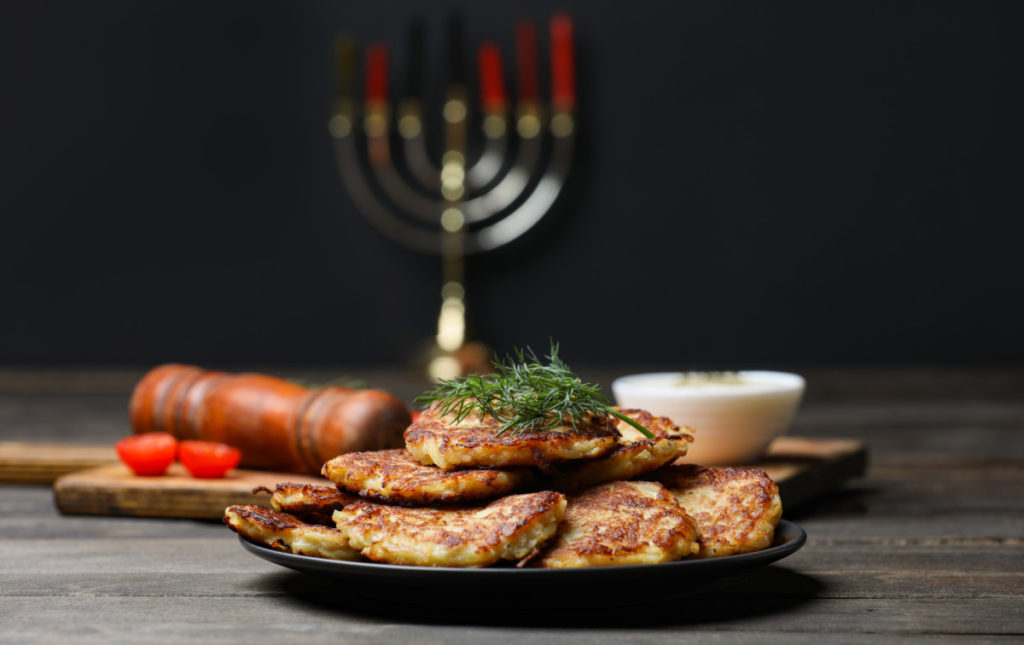 A plate of latkes on a dark wood tabletop, behind there is a menorah on a cutting board, a pepper grinder and a sliced tomato. The background is dark. 