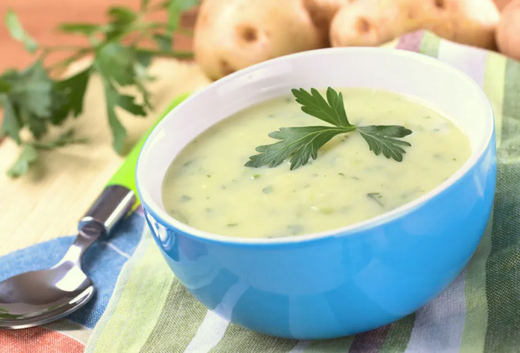 A bowl of creamy potato and parsley soup set on a striped napkin. There are potatoes in the background.