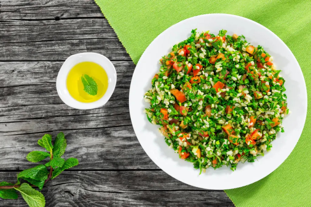 Overhead view of a white bowl filled with tabbouleh. The bowl sits on a lime green tablecoth. There is a sprig of mint next to the bowl. 