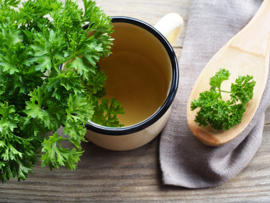 A cup of parsley tea in a ceramic mug. There is a decorative napkin with a wood spoon next to the mug. A large bunch of parsley is to the left.