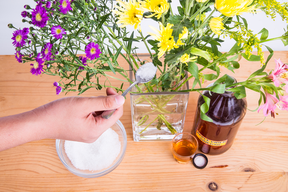 a hand holds a spoonful of sugar above a vase full of flowers. Next to the vase is a bottle of apple cider vinegar.