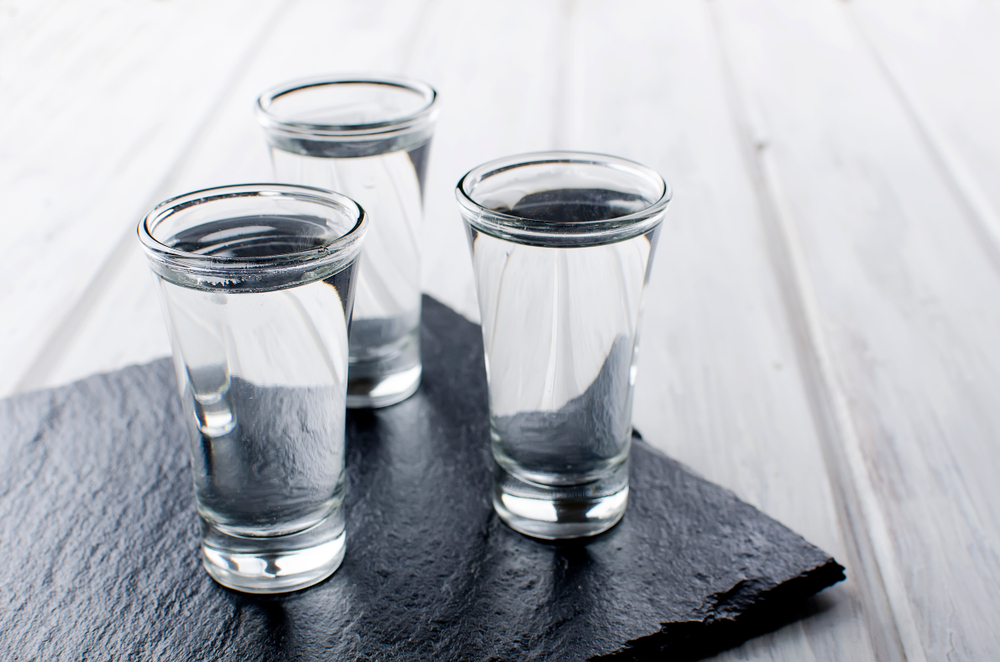 Three shots of vodka in clear shot glasses. The glasses are sitting on a slab of slate on a white wood table.