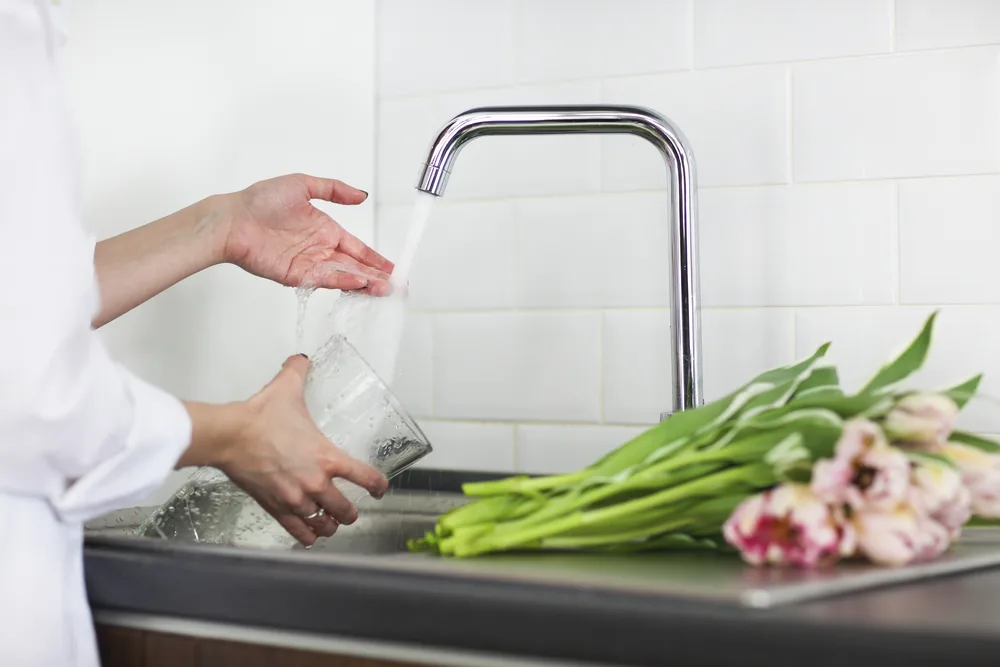 A woman stands at a sink with her hand under the running water. She is holding an empty vase in the other hand. There are pink tulips on the counter next to her.