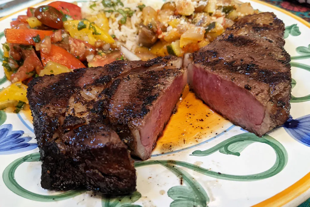 A dinner plate with a dry-aged ribeye steak cut open to reveal a pink inside. There is a vegetable dish on the other side of the plate.