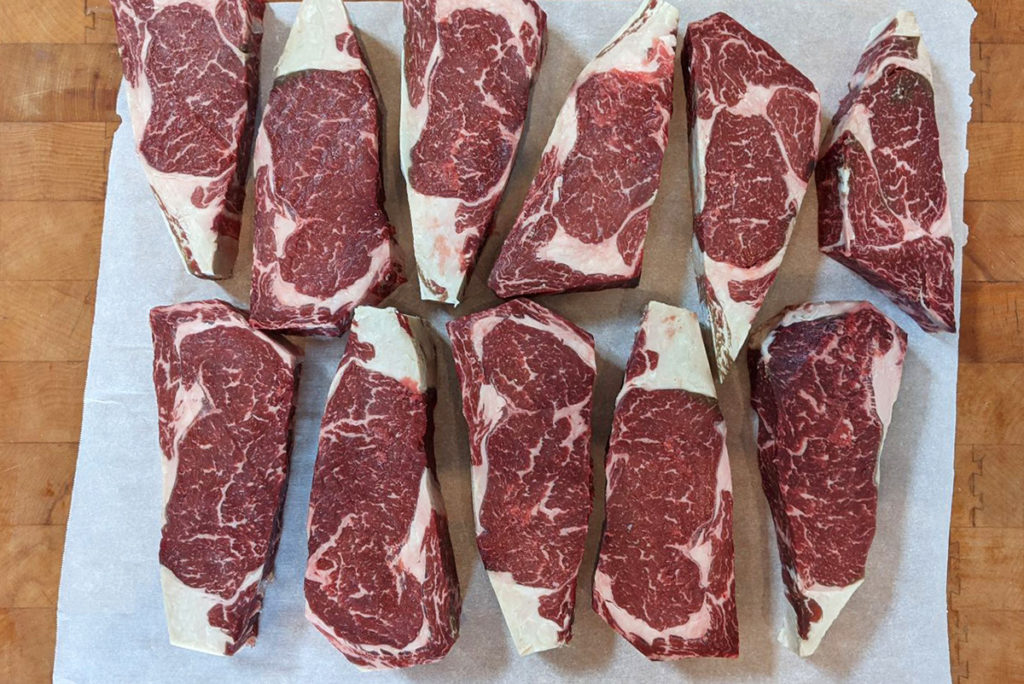 Overhead view of dry-aged ribeye steaks trimmed of bark laying on a piece of parchment paper on a butcher block. 