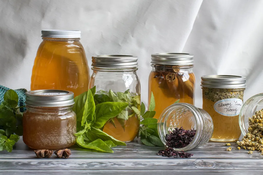 Several jars of honey with herbs infusing in them. There is a jar of honey in the back and fresh and dried herbs scattered in front of the other jars. 