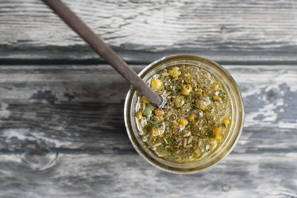 Overhead view of a jar filled with honey, chamomile flowers, lavender buds, and dried peppermint. A chopstick is sticking up out of the jar. 