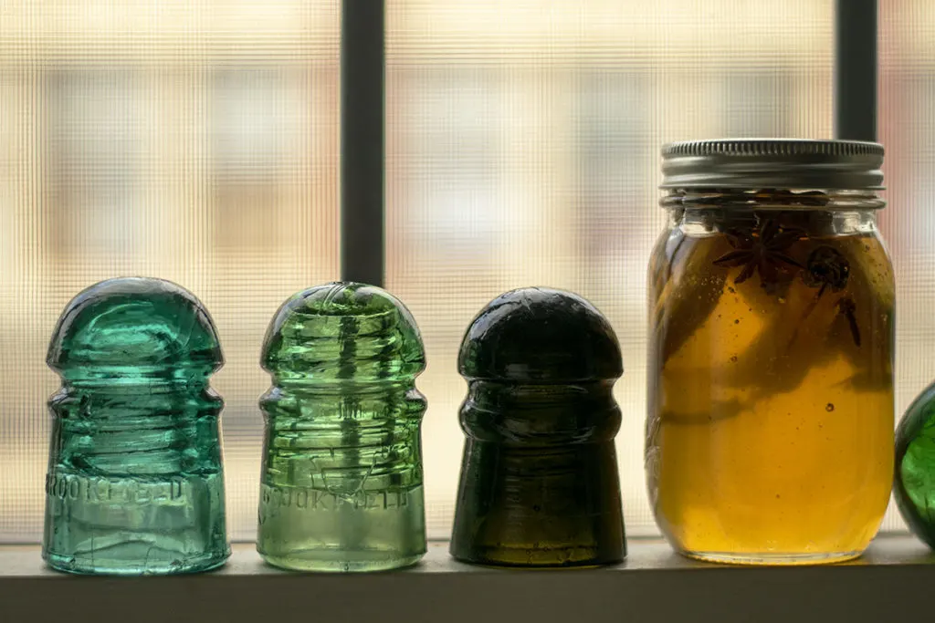 View of a windowsill with glass insulators on it and a jar of honey with cinnamon sticks star anise and cloves floating in it.