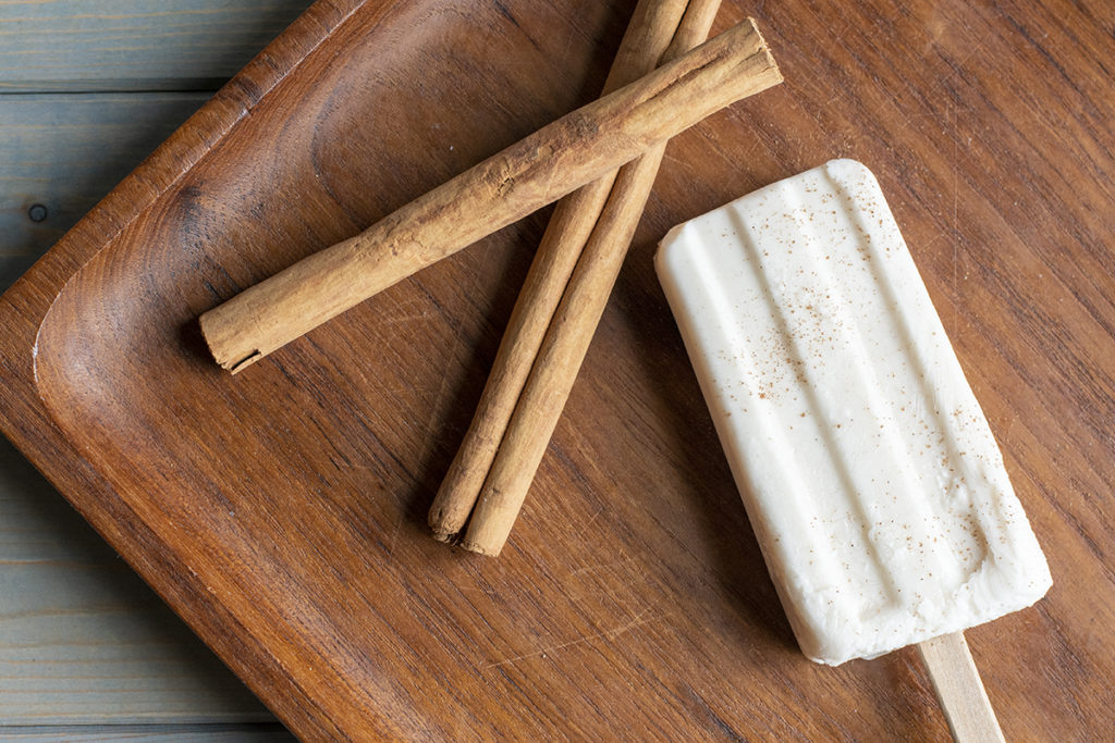 Cream colored horchata popsicle sprinkled with ground cinnamon next to cinnamon sticks