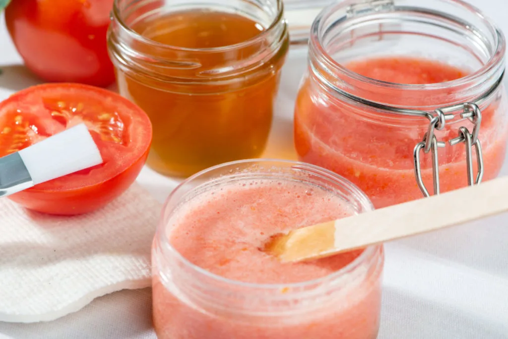 A jar of frothy tomato puree next to a jar of honey and a sliced tomato. There is a small paint brush resting on the tomato and a wooden popsicle stick in the homemade face mask