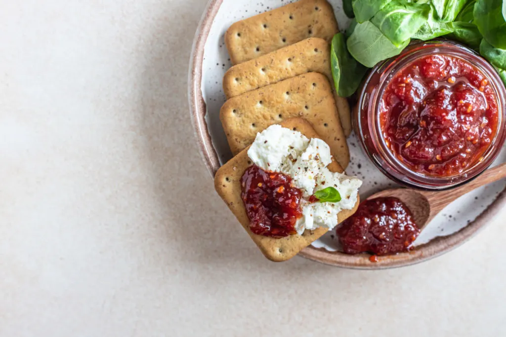 A jar of tomato jam with crackers and goat cheese and tomato jam spread on them
