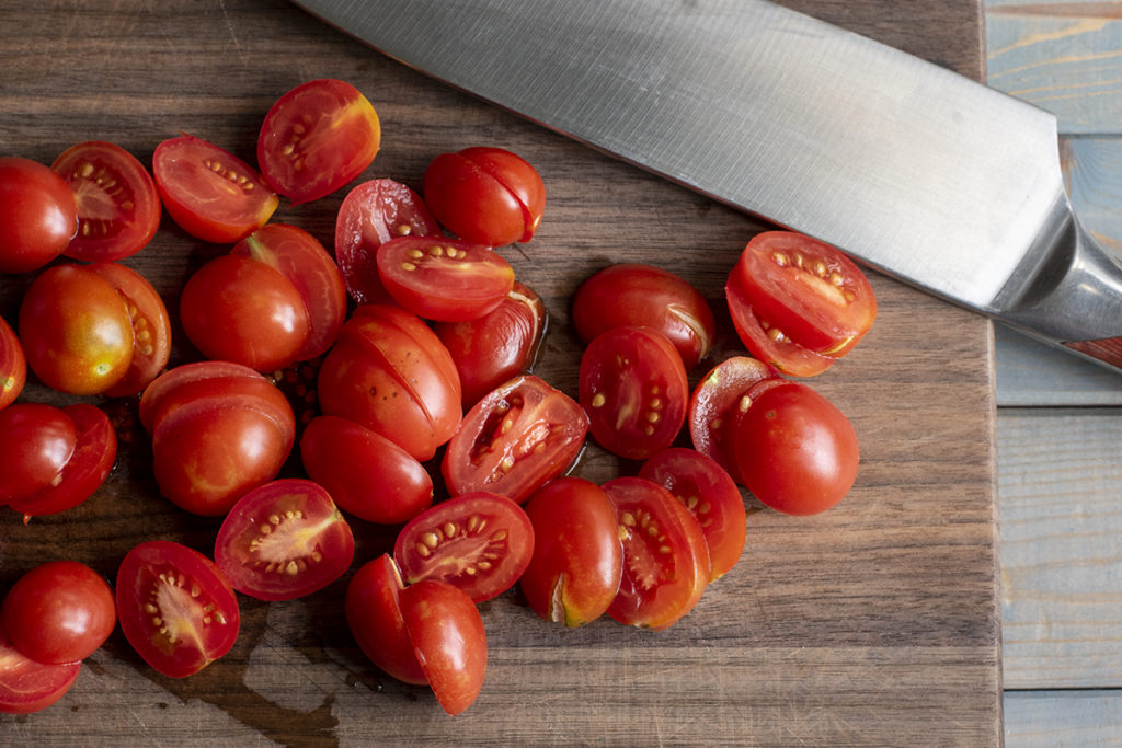 A cutting board covered in halved cherry tomatoes. There is a chefs knife at the top of the cutting board.