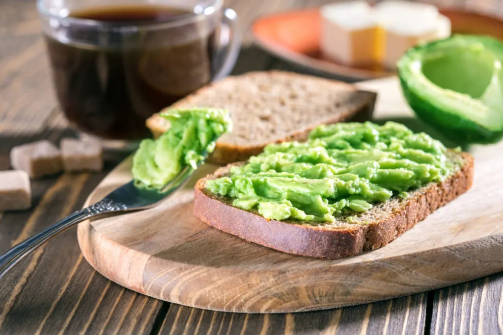 Whole wheat bread with avocado spread on it, there is a cup of coffee in the background. 