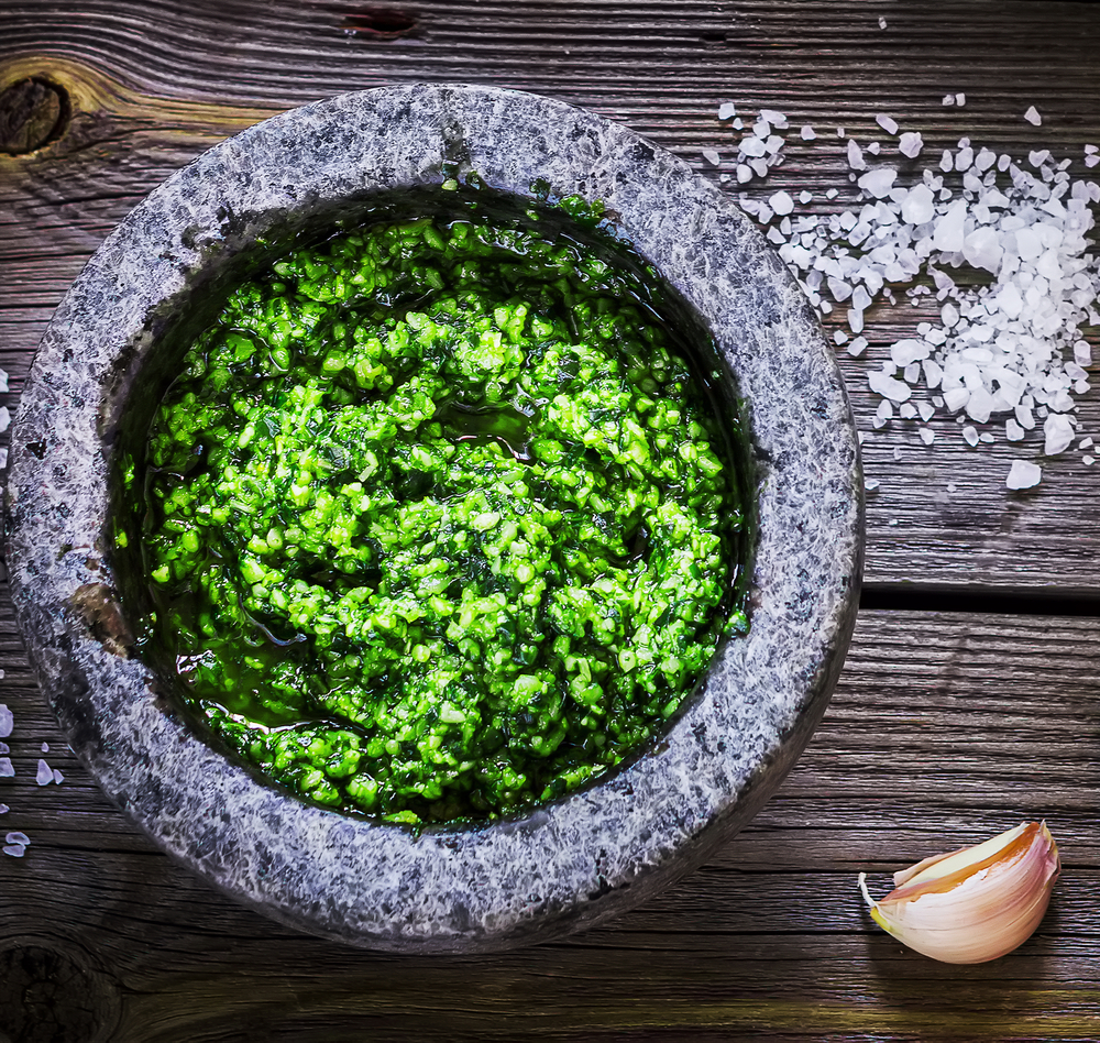 Overhead view of a marble mortar filled with freshly ground chive pesto. There are chunks of sea salt on the rough wood table next to it. 