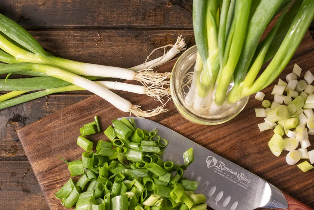 A cutting board with a knife and chopped green onions. There are whole green onions standing upright in a jar of water and several green onion laying next to the cutting board.