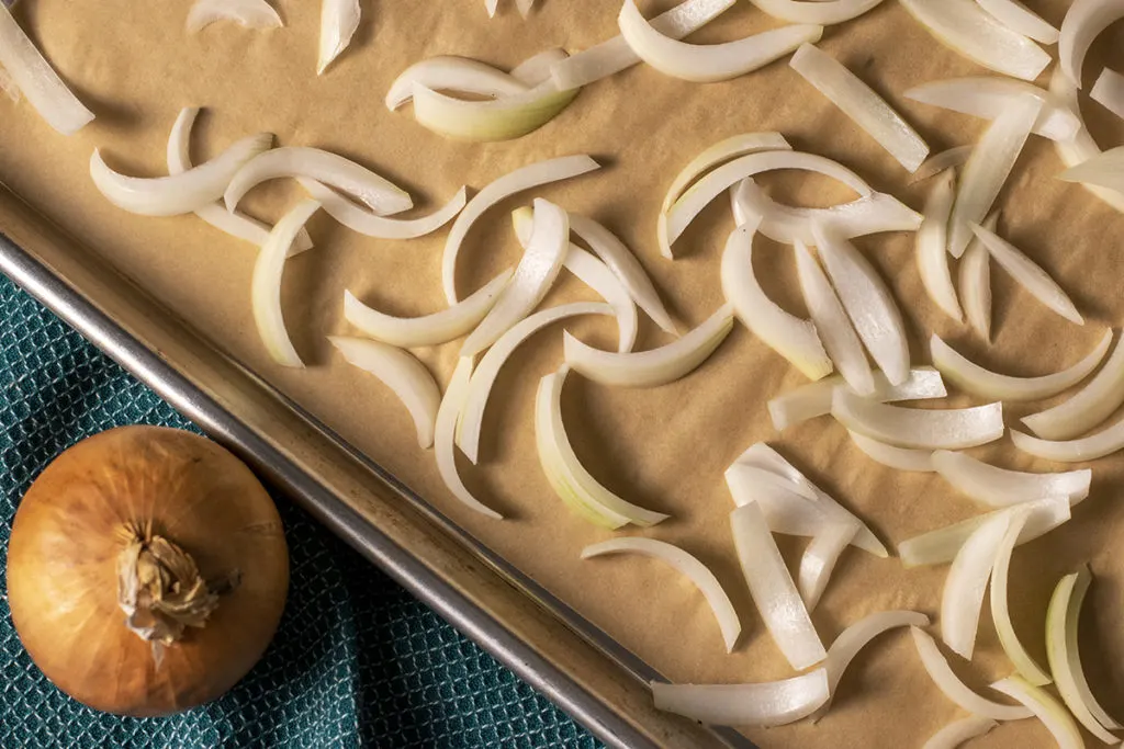 A parchment-lined baking sheet with slivered onions spread across it. 