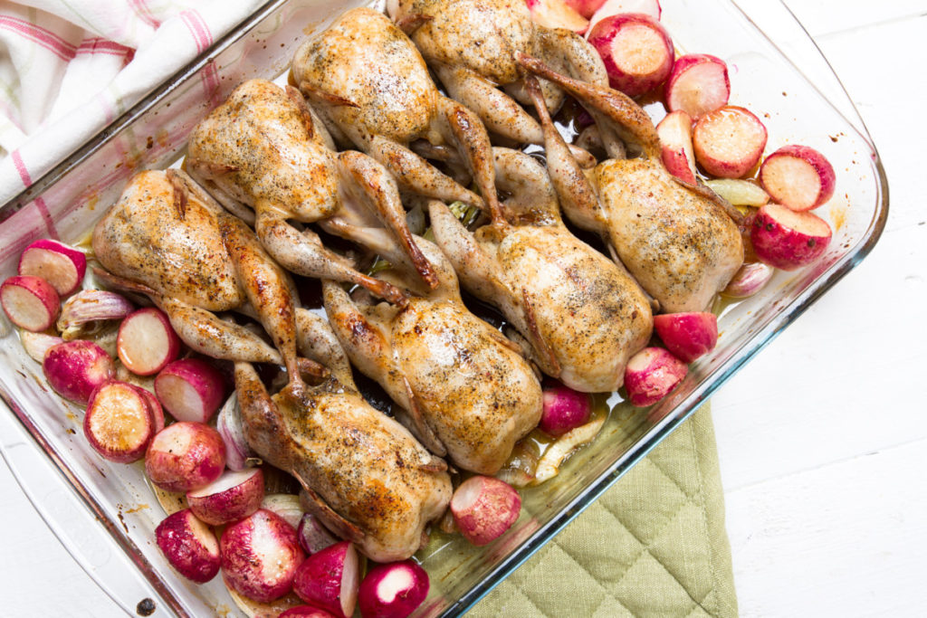 A clear glass baking dish holds eight roasted quail surrounded by roasted radishes.