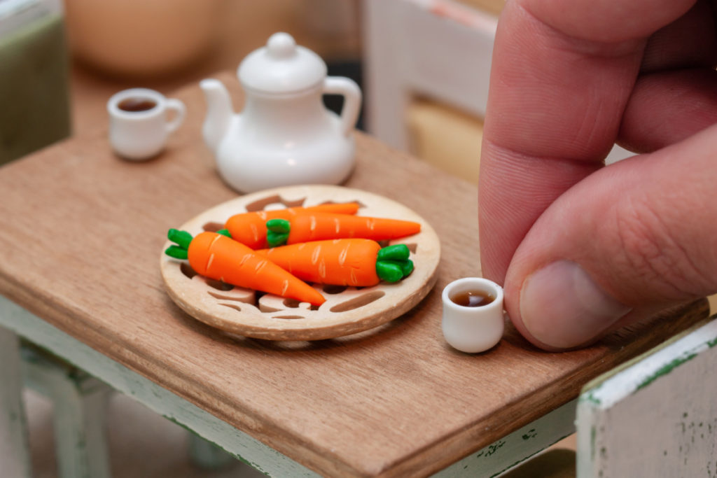 Someone's fingers setting down a tiny cup of coffee on a dollhouse table with tiny carrots on it. 
