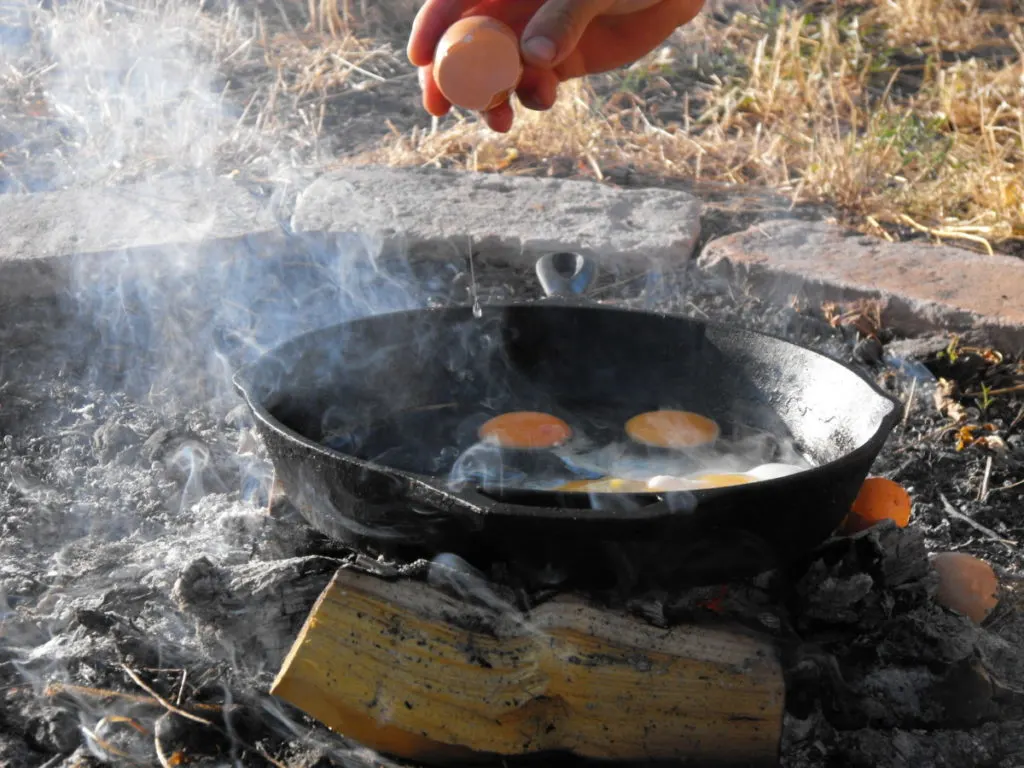 Close up of a hand cracking an egg into a cast iron frying pan set over a campfire. 