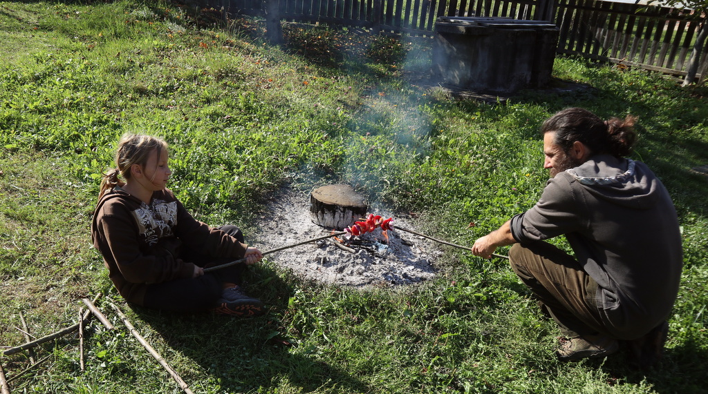 A father and daughter sit by a campfire cooking food on sticks. 