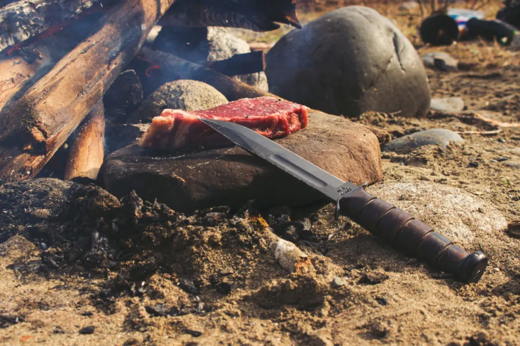 A steak being prepared to cook over a campfire. The steak is sitting on a rock with a large hunting knife next to it.
