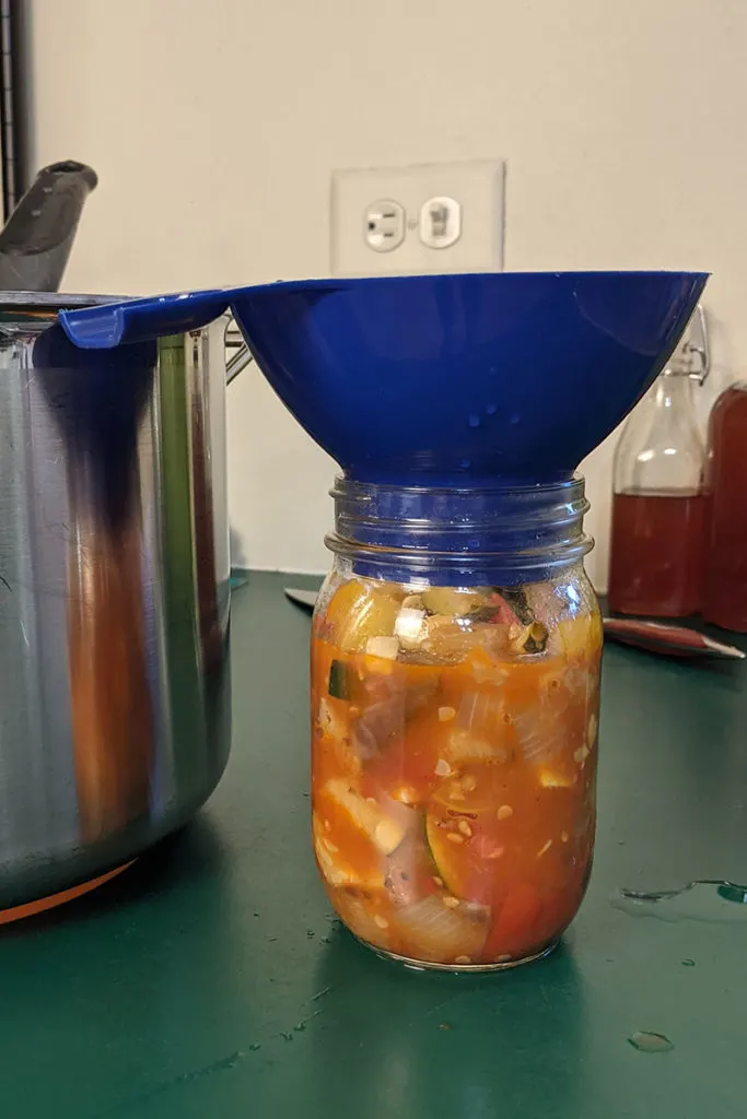 A jar filled with hot ratatouille with a canning funnel put inside it.