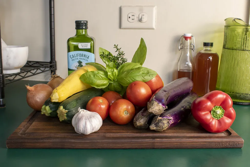 A large cutting board with the ingredients for ratatouille stacked on it. Olive oil, garlic, onions, summer squash, tomatoes, eggplants, peppers and sprigs of basil and thyme. 
