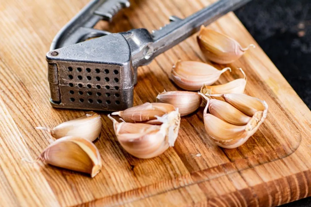 A metal garlic press laying next to garlic cloves on a cutting board.