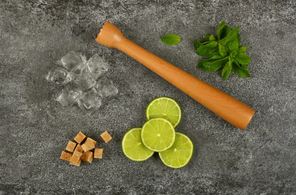 A wooden muddler next to ice cubes, mint, lime slices, and demerara sugar cubes.