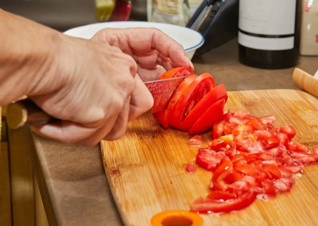 A man's hands are shown. He is holding and slicing a tomato on a cutting board.