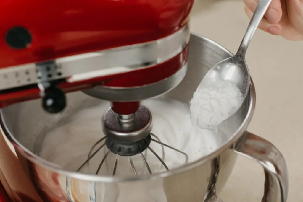 Red Kitchen aid stand mixer with whipped egg whites in the bowl. A hand holding a spoon above it.