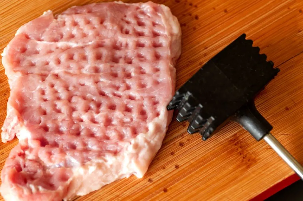 A plastic meat tenderizer laying next to a pulverized pork chop.