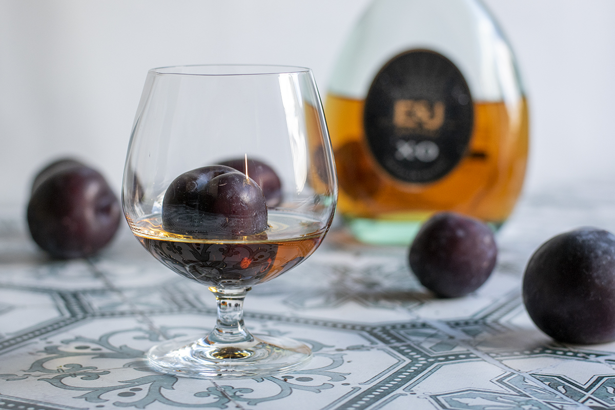A brandy snifter with a plum sitting in brandy. Bottle of brandy and plums in soft focus in the background. 