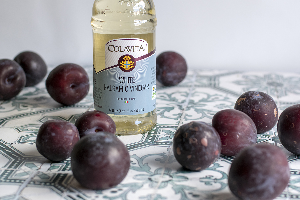 A bottle of white balsamic vinegar surrounded by purple plums