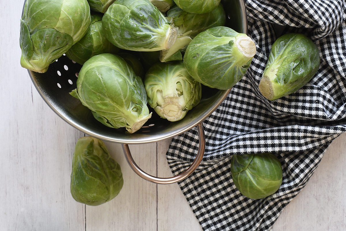 Brass colander with freshly washed Brussels sprouts next to a black and white checkered dishtowel.