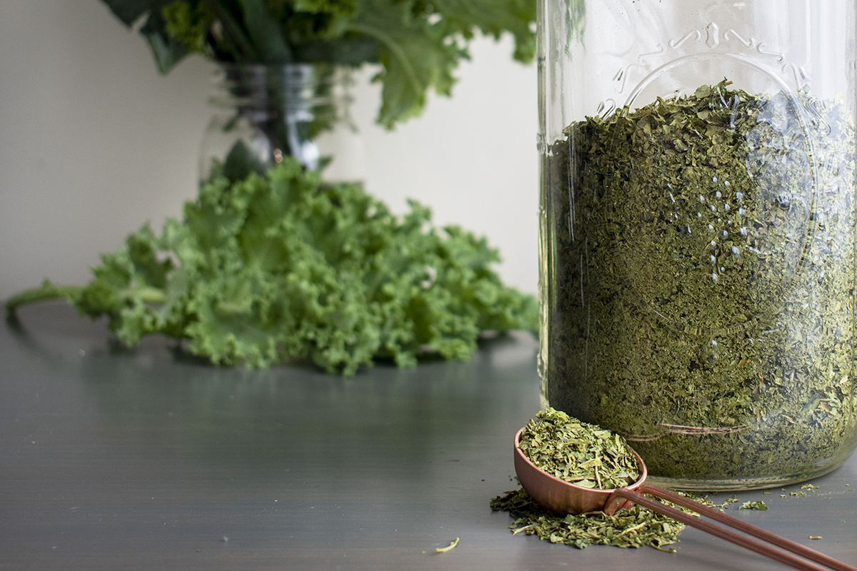 Jar of dried smoothie greens with measuring spoon and fresh kale in soft focus in the background