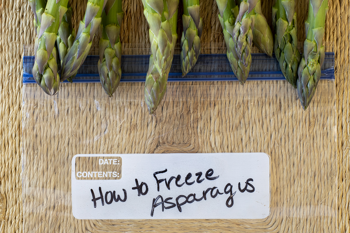 Asparagus stems lined up along the top of a ziptop freezer bag with 'How to Freeze Asparagus' written on it.