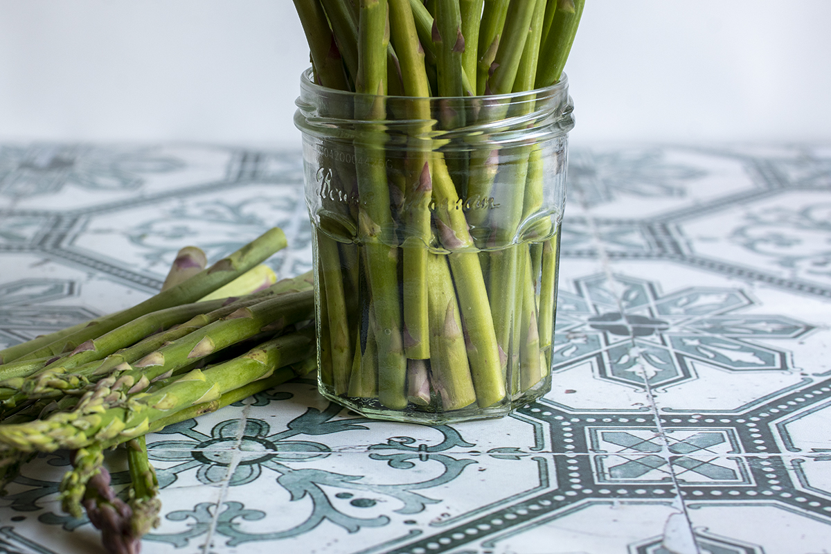 Jar with water and asparagus stalks in it, stalks lying next to the jar. 