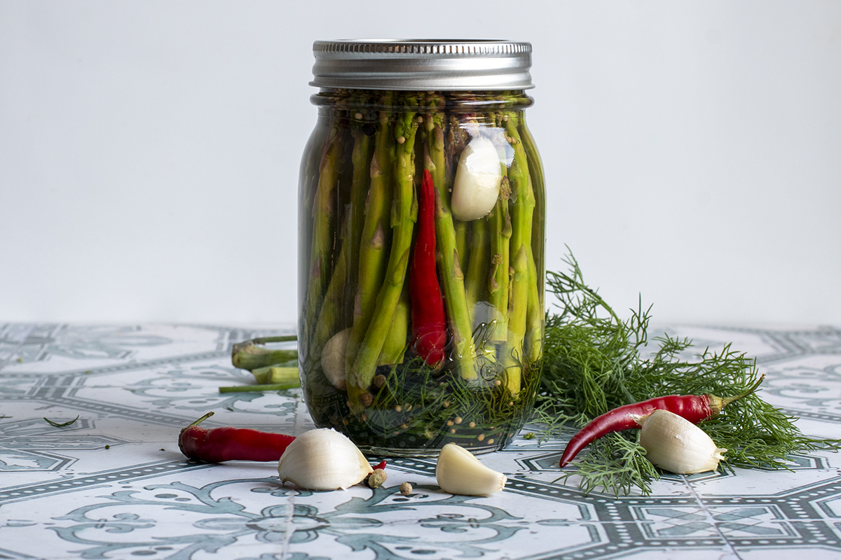 A jar of quick dilly asparagus pickles, surrounded by fresh dill, peppercorns, garlic cloves and hot peppers. 