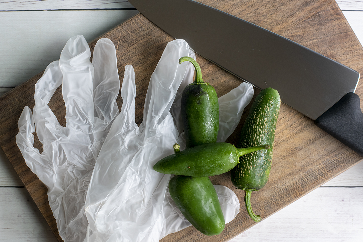 Overhead view of fresh jalapenos, vinyl gloves and a chefs knife on a cutting board.
