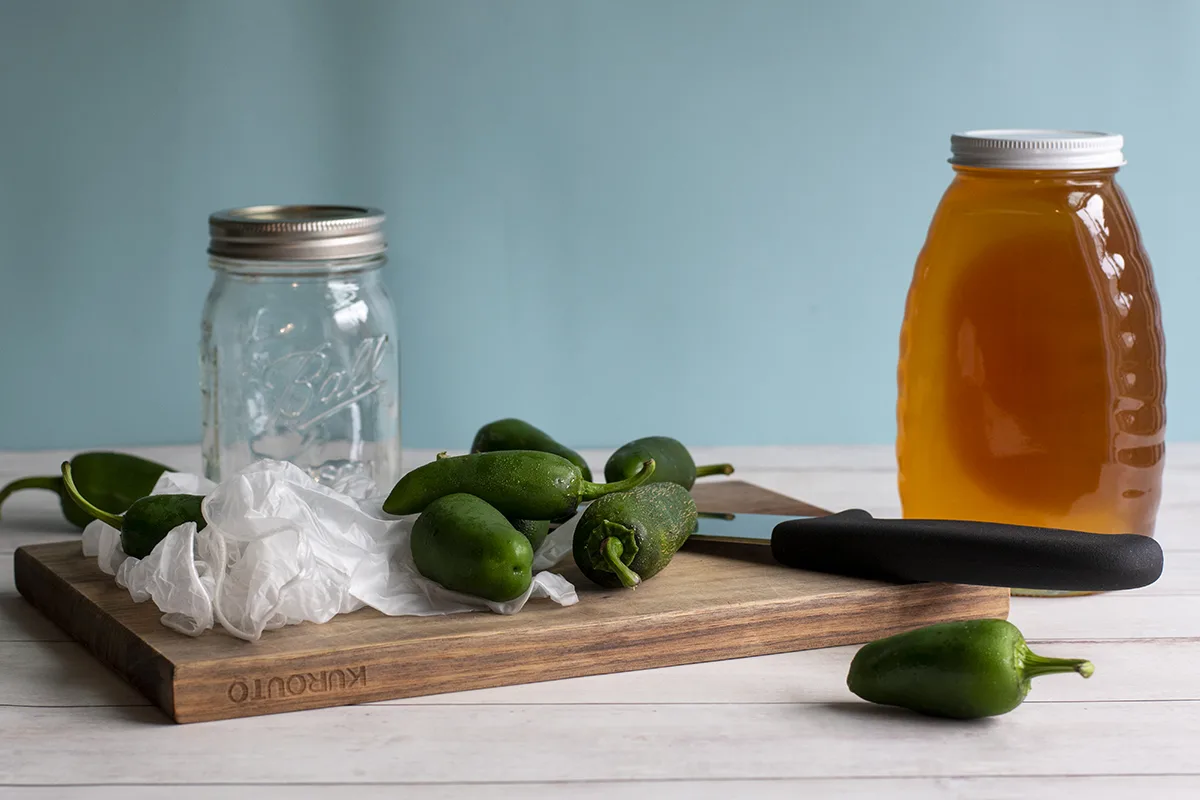 Cutting board, jar of honey, empty mason jar, disposable gloves, chefs knife and fresh jalapenos